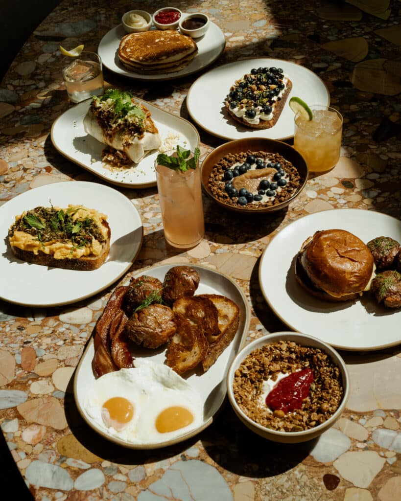 Overhead view of a brunch spread on a stone-inlay table, with eggs, bacon, and potatoes; pancakes; scrambled egg toast; a blueberry tart; a burrito; a burger; granola bowls; and cocktails.