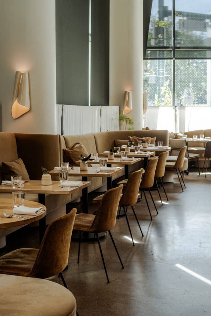 Row of wooden tables set with napkins and small plants along a tan velvet banquette, beneath sculptural wall sconces and tall windows.