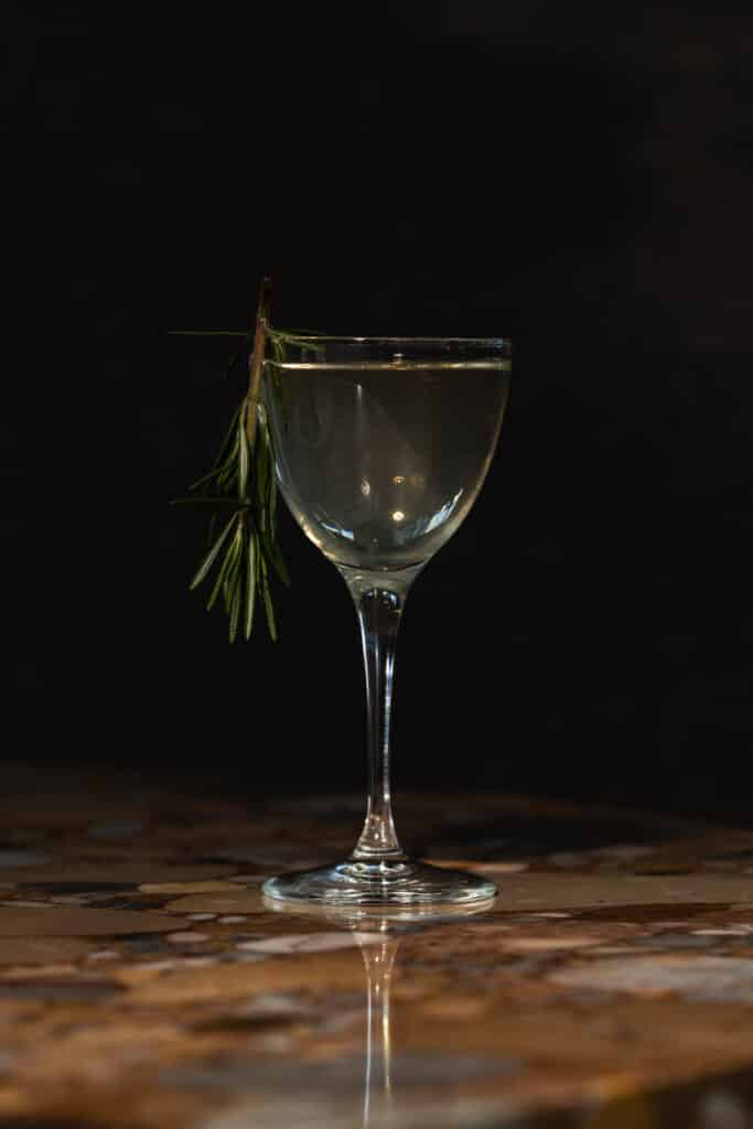 Clear cocktail in a stemmed glass garnished with a sprig of rosemary, on a stone-inlay table against a dark background.