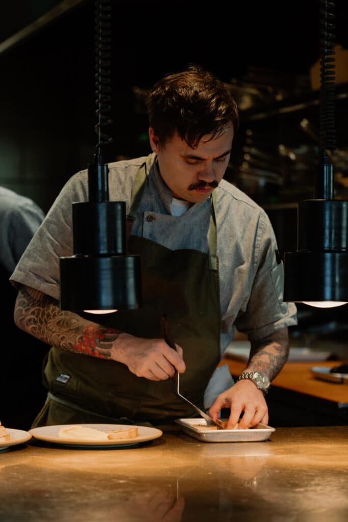 Chef with tattooed forearms plating food under black heat lamps at a polished kitchen counter.
