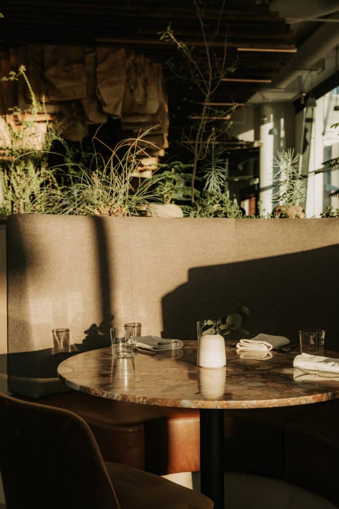Round stone-inlay table set for dinner in a gray booth, backed by a planter of grasses and greenery in late-afternoon light.